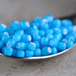 Puede incluir: Una cuchara llena de peque&ntilde;as cuentas de vidrio azul claro con un centro blanco. Las cuentas son redondas y tienen un acabado brillante.