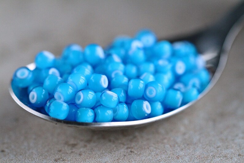 Puede incluir: Una cuchara llena de peque&ntilde;as cuentas de vidrio azul claro con un centro blanco. Las cuentas son redondas y tienen un acabado brillante.