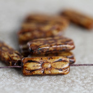 Könnte beinhalten: Nahaufnahme einer Gruppe brauner und beigefarbener rechteckiger Keramikperlen mit einem geschnitzten Design. Die Perlen sind auf einer dünnen braunen Schnur aufgefädelt.