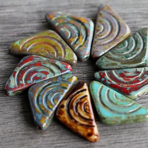 May include: A collection of ten triangular ceramic beads with a swirling, multi-colored pattern. The beads are arranged in a circular pattern on a wooden surface.