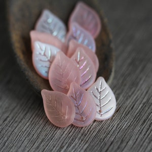 May include: A group of pink glass leaf-shaped beads with a silver leaf design. The beads are arranged on a wooden surface.