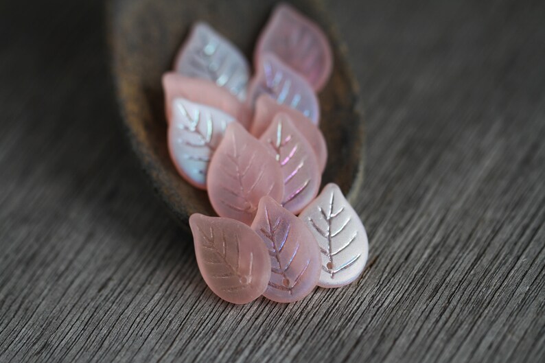 May include: A group of pink glass leaf-shaped beads with a silver leaf design. The beads are arranged on a wooden surface.