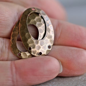 K&ouml;nnte beinhalten: Zwei ovale Metall-Anh&auml;nger mit geh&auml;mmerter Textur und einem ausgeschnittenen Oval in der Mitte. Die Anh&auml;nger haben jeweils drei L&ouml;cher zum Anbringen an Schmuck oder anderen Bastelarbeiten.