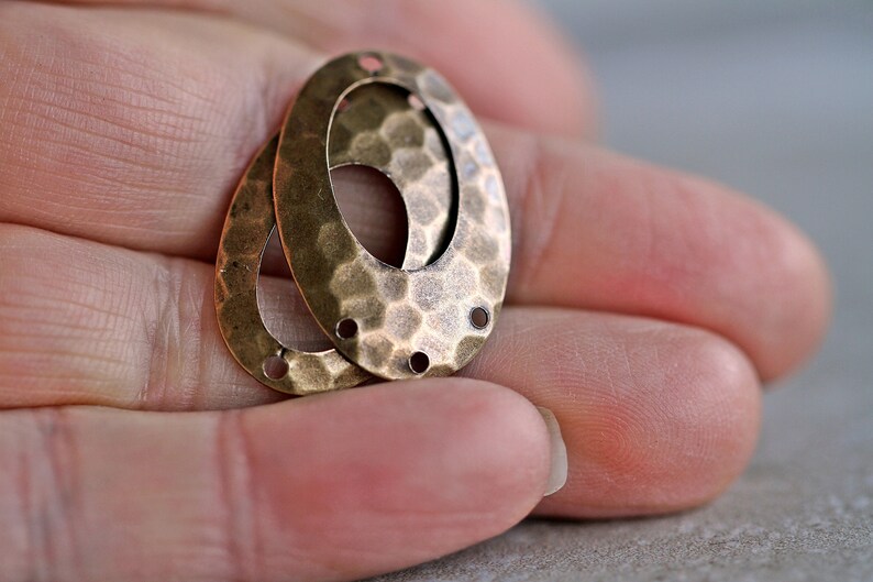 K&ouml;nnte beinhalten: Zwei ovale Metall-Anh&auml;nger mit geh&auml;mmerter Textur und einem ausgeschnittenen Oval in der Mitte. Die Anh&auml;nger haben jeweils drei L&ouml;cher zum Anbringen an Schmuck oder anderen Bastelarbeiten.