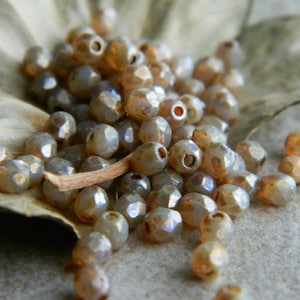 May include: A close-up of a pile of iridescent glass beads in shades of beige and brown. The beads are faceted and have a shimmery finish.