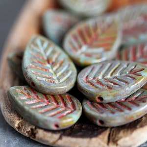 May include: A collection of teal and red leaf-shaped ceramic beads with a textured surface. The beads are arranged in a wooden bowl.