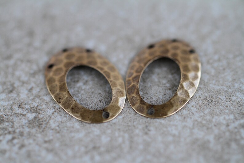 K&ouml;nnte beinhalten: Zwei ovale Metall-Anh&auml;nger mit geh&auml;mmerter Textur. Die Anh&auml;nger sind hellgoldfarben und haben oben ein Loch zum Anbringen an Schmuck.