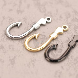 May include: Three metal fish hook charms in silver, gold, and black. Each hook has a curved point and a detailed handle with a loop for attaching to jewelry or other crafts. The hooks are displayed on a textured gray surface.