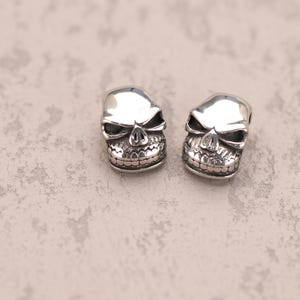 Könnte beinhalten: Zwei silberne Totenkopfperlen. Jede Perle hat ein detailliertes Totenkopfdesign mit hohlen Augenhöhlen, einer markanten Nase und einem detaillierten Kiefer. Die Schädel haben eine polierte Oberfläche, die das Licht reflektiert. Die Perlen sind wahrscheinlich für die Schmuckherstellung.