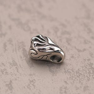 Puede incluir: Un colgante de metal plateado con un diseño intrincado y en espiral. El colgante tiene un agujero en la parte superior para sujetarlo a una cadena o cordón.