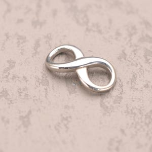 Puede incluir: Un dije de símbolo de infinito de plata pulida. El dije tiene un diseño suave y redondeado, con forma de ocho. El fondo es una superficie texturizada de tono neutro.