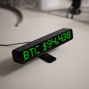 May include: A black digital display showing "BTC $94,438" in bright green LED lights. The rectangular device is angled on a black stand, with a textured surface. A black cable extends from the device.