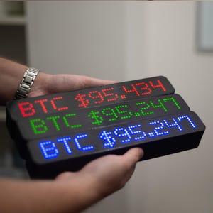 Könnte beinhalten: Drei schwarze LED-Anzeigen mit Bitcoin-Preisen in Rot, Grün und Blau. Jede Anzeige zeigt "BTC" gefolgt von einem Dollarbetrag. Die Anzeigen werden in den Händen einer Person gehalten, mit einer silbernen Uhr am Handgelenk.