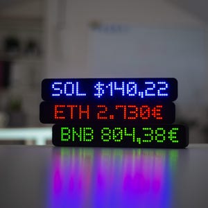 Może przedstawiać: Trzy czarne wyświetlacze LED ułożone jeden na drugim, pokazujące dane kryptowalut. Górny wyświetlacz pokazuje "SOL $140.22" w kolorze niebieskim. Środkowy wyświetlacz pokazuje "ETH 2.730€" w kolorze czerwonym. Dolny wyświetlacz pokazuje "BNB 804,38€" w kolorze zielonym.