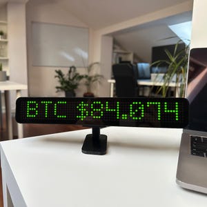 Może przedstawiać: Czarny wyświetlacz cyfrowy z napisem "BTC $84,074" w zielonych diodach LED. Wyświetlacz jest zamontowany na małym czarnym stojaku i stoi na białej powierzchni. Częściowo widoczny po prawej stronie laptop.