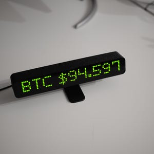 May include: A black digital display showing "BTC $94.597" in green LED lights. The display is rectangular and sits on a small black stand. A black cable extends from the display.