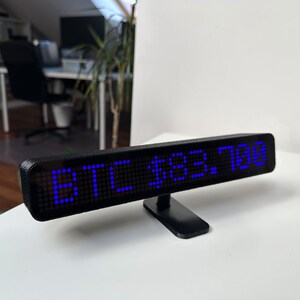 Może przedstawiać: Czarny wyświetlacz LED z napisem "BTC $83.700" w jasnoniebieskim kolorze. Wyświetlacz jest prostokątny i zamontowany na małym czarnym stojaku. Tło to biała powierzchnia z rozmytym otoczeniem biurowym.