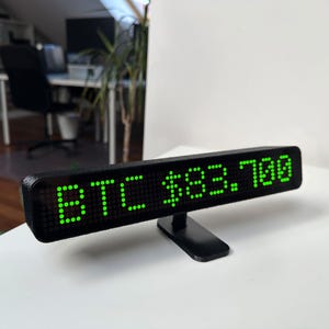 May include: Black LED display showing "BTC $83,700" in bright green dots. The display is mounted on a black stand. The background is a white surface with a blurred office setting.