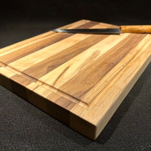 May include: A rectangular wooden cutting board with a juice groove and a knife. The board is made of light and dark wood strips. The knife has a wooden handle and a silver blade. The cutting board is approximately 12 inches wide and 16 inches long.