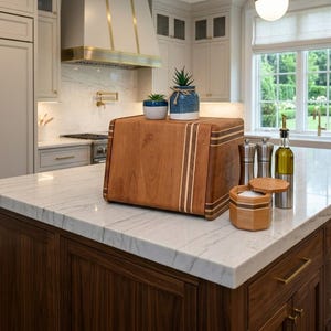May include: A rectangular wooden bread box with a smooth finish and light-colored vertical wood inlay stripes. Two small potted succulents sit on top. Next to the box are two metal salt and pepper grinders and a glass bottle of olive oil.