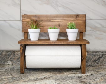 May include: A dark brown wooden paper towel holder featuring a top shelf with three small white pots containing green artificial plants. The holder has a horizontal rod underneath the shelf that supports a roll of white paper towels.