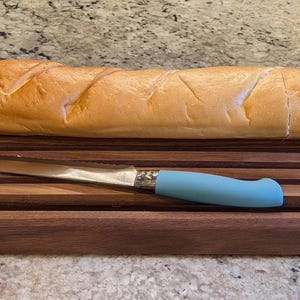May include: A rectangular dark brown wooden bread board with horizontal slats, featuring a long, golden-brown baguette sliced into pieces and a serrated bread knife with a light blue handle resting on the surface of the board.