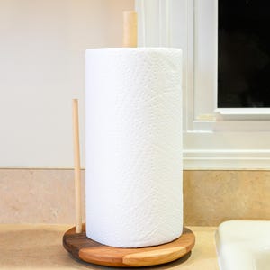 May include: A wooden paper towel holder with a roll of white paper towels. The holder has a round base with a two-tone wood design and a wooden dowel to hold the roll in place. The holder is on a beige countertop.