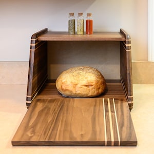 May include: A dark wood bread box with a drop-down front door featuring light wood inlay stripes. Inside, a round loaf of bread sits on the base, while three small glass spice jars with cork stoppers are placed on the top shelf.