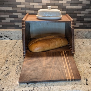 May include: A dark brown wooden bread box with a drop-down front door featuring light wood inlay stripes. Inside, a loaf of golden-brown bread sits on the base. A white ceramic butter dish with a lid rests on the top surface of the bread box.