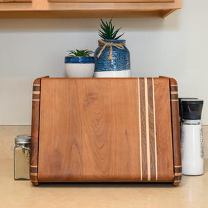 May include: A rectangular wooden cutting board with rounded edges and decorative stripes. The board is made of light and dark brown wood. Two small potted plants and a salt and pepper shaker are on the counter.