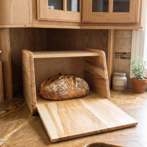 May include: A light-colored wooden bread box with a drop-down front door that serves as a shelf. Inside the box sits a single round loaf of crusty, golden-brown sourdough bread with a scored top, showcasing natural wood grain patterns.