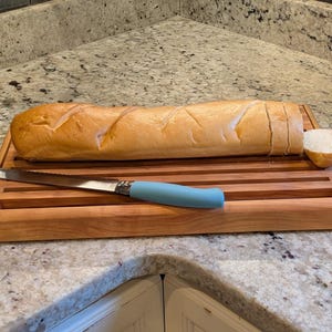 May include: A rectangular wooden bread board with horizontal slats and a light brown finish. A long, golden-brown baguette is sliced on the board, accompanied by a serrated bread knife featuring a smooth, light blue handle resting on the surface.