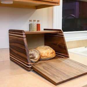 May include: A wooden bread box with a hinged lid, crafted from dark brown wood with light wood accents. The box is open, revealing two loaves of bread. Three small glass spice jars sit on a shelf above.