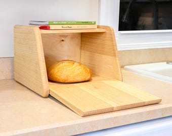 May include: A light-colored wooden bread box with a drop-down front door, containing a round loaf of crusty bread. On top of the bread box sits a green book titled short and simple and a tan book titled Buca di Beppo, both with visible text.
