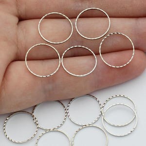 May include: A collection of small, silver-colored metal rings, some with a textured, twisted design. The rings are scattered on a light background, suggesting they are craft or jewelry components.