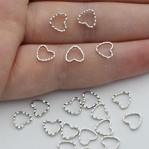 May include: Small, silver-colored heart-shaped charms. Some charms have a textured edge, while others have a smooth edge. The charms are scattered on a white surface, with some resting on a person's hand.