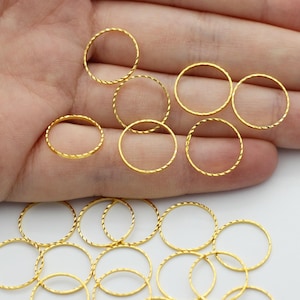 May include: A collection of small, gold-colored metal rings, some with a textured, twisted design. The rings are scattered on a white surface, with some held in an open hand. The rings are likely jewelry findings or components.