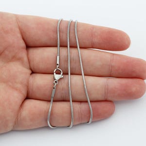 May include: A silver-colored snake chain necklace with a lobster clasp. The chain is smooth and flexible, and it is displayed on a light background.