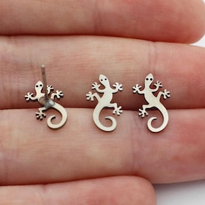 May include: Silver-colored lizard stud earrings and a single earring post. The earrings are small, with a detailed lizard design featuring a curled tail. The earrings are displayed on a light background.