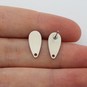 May include: A pair of silver-colored teardrop-shaped stud earrings. Each earring has a small hole at the bottom and a post on the back for securing. The earrings are displayed on a light background.