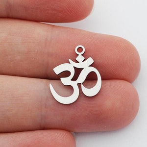 Puede incluir: Dos amuletos de plata con el símbolo Om y un agujero para colgar. Los amuletos están sobre un fondo blanco.
