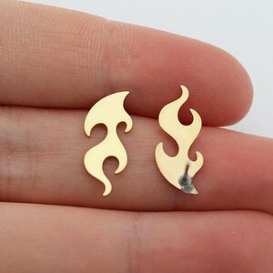 Puede incluir: Dos pendientes dorados con forma de llama. Los pendientes son pequeños, con una superficie lisa y pulida. El diseño de la llama tiene bordes curvos y una parte superior puntiaguda, creando un aspecto dinámico. Se muestran sobre un fondo claro.