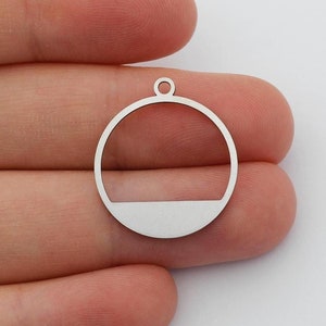 May include: A silver-colored metal charm with a circular design. The charm features a partial circle with a flat bottom section and a small loop at the top for attaching to a chain or other jewelry component. The charm is held in a hand.