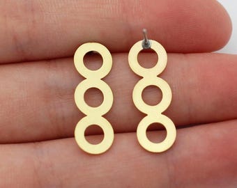 Puede incluir: Un par de pendientes dorados, cada uno con tres formas circulares conectadas en línea vertical. Los pendientes se muestran sobre un fondo neutro, destacando su diseño simple y moderno. Se ve el poste de un pendiente.
