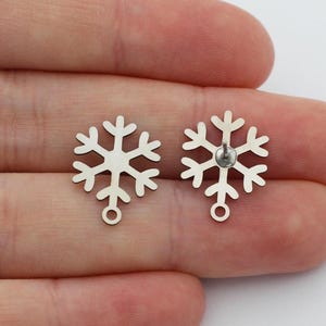 May include: Two silver snowflake charms with a loop at the bottom for jewelry making. One charm has a small silver post on the back. The charms are displayed on a light background.