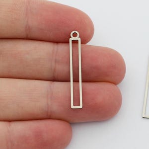 May include: Two silver-colored, rectangular charms with a small loop at the top for jewelry making. One charm is held in a hand, while the other lies separately. The charms have a simple, minimalist design.