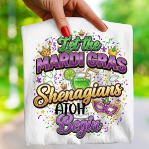 Puede incluir: Camiseta blanca con un diseño colorido de Mardi Gras. El diseño incluye el texto "Let the Mardi Gras Shenanigans Begin" en verde, morado y dorado, junto con un cóctel, una corona y una máscara.
