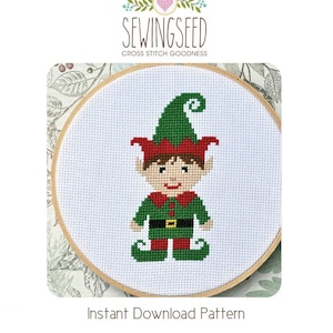 May include: Cross stitch pattern of a green elf with a red hat and a black belt. The elf is smiling and has a brown hair. The pattern is in a white embroidery hoop.