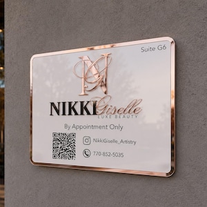 May include: A rectangular white sign with a rose gold metallic border and trim. The sign features the text NIKKI Giselle LUXE BEAUTY, Suite G6, By Appointment Only, a QR code, an Instagram icon with NikkiGiselle_Artistry, and a phone icon with 770-852-5035.