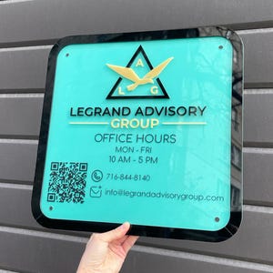 May include: A square sign with a teal background and black trim. The sign features the text "LEGRAND ADVISORY GROUP" and "OFFICE HOURS MON-FRI 10 AM - 5 PM". A gold logo and contact information are also displayed.